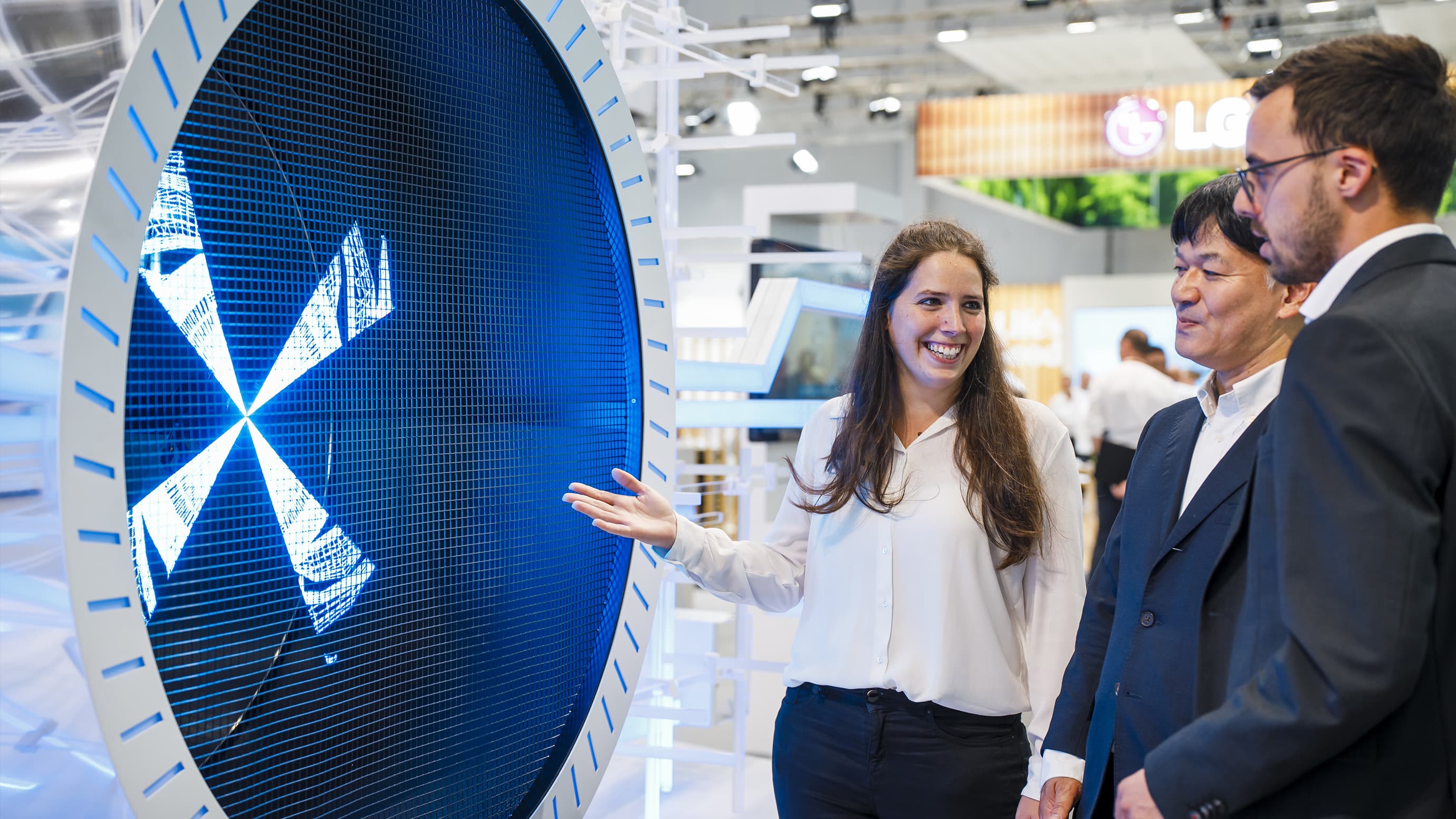 A female exhibitor stands in front of a fan with two other trade fair participants and presents the product